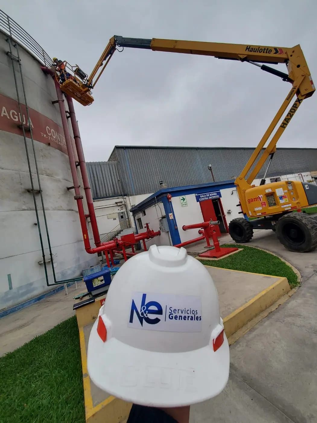 Manlift posicionado para trabajo en altura junto a tuberías en planta industrial, casco de N&E en primer plano